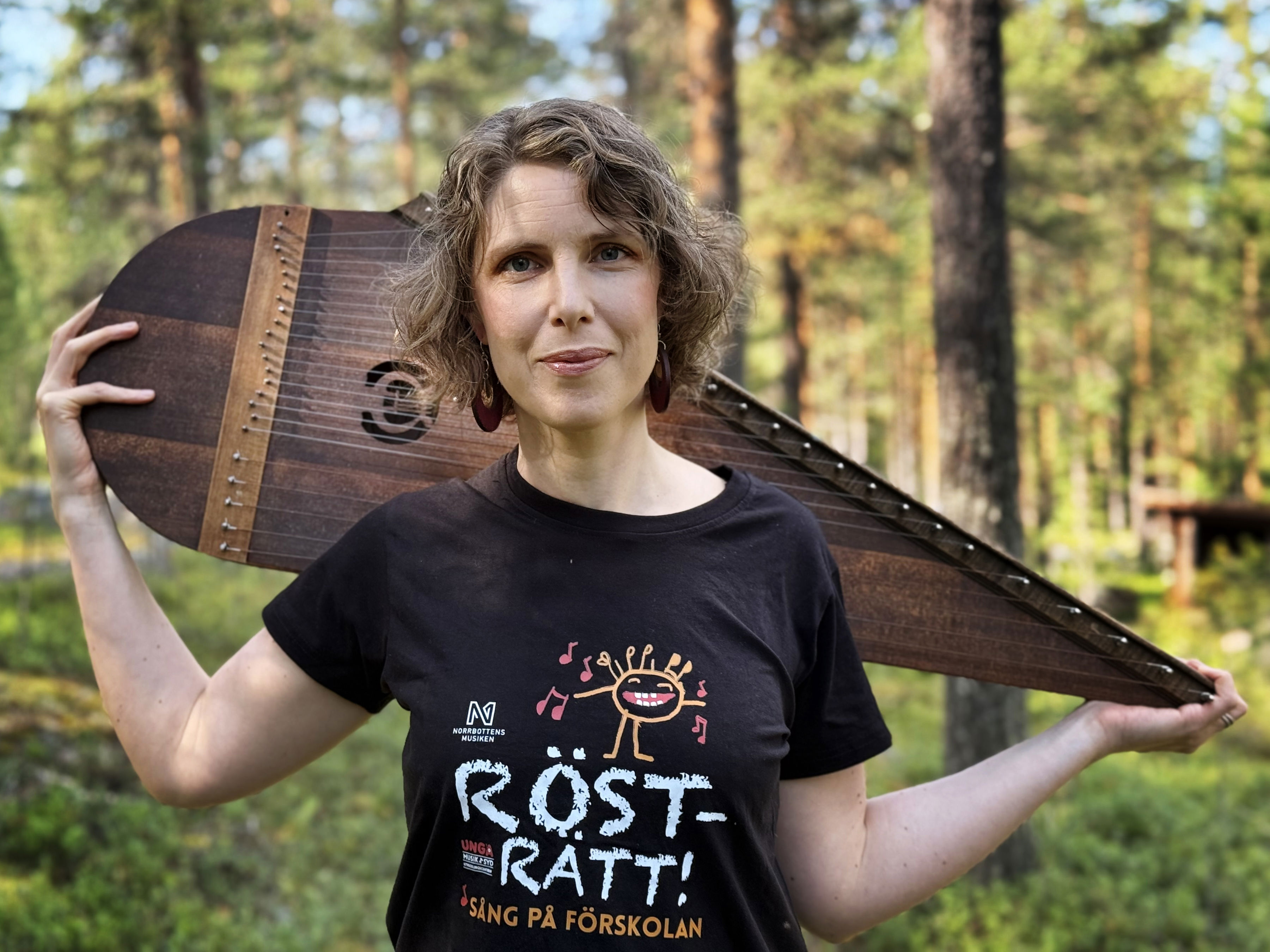 Foto av Mirja Palo som står i skogen och håller sitt instrument - en kantele.